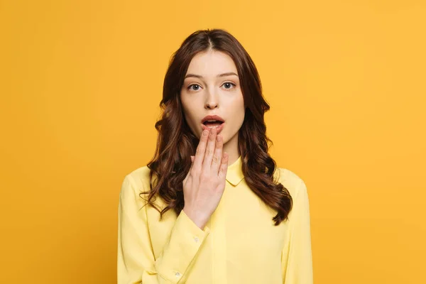 Chocada chica sosteniendo la mano cerca de la boca abierta mientras mira a la cámara aislada en amarillo - foto de stock