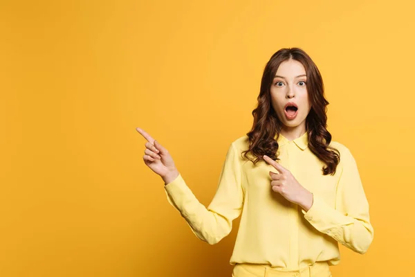 Menina chocada apontando com os dedos enquanto olha para a câmera com a boca aberta no fundo amarelo — Fotografia de Stock