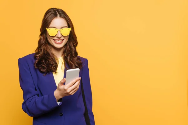 Cheerful, stylish girl in yellow glasses chatting on smartphone isolated on yellow — Stock Photo