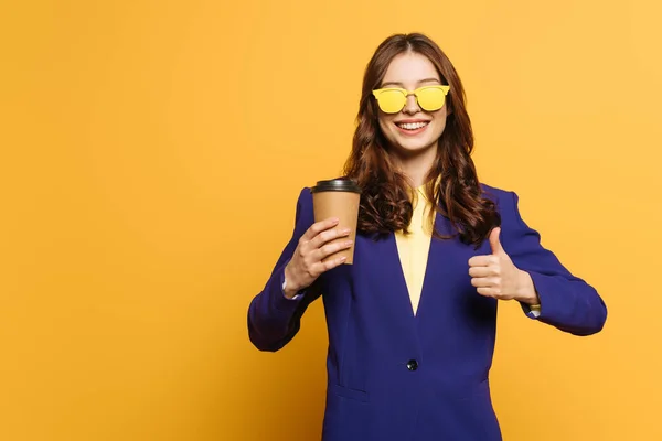 Chica elegante y sonriente en gafas amarillas sosteniendo café para ir y mostrando el pulgar hacia arriba sobre fondo amarillo - foto de stock