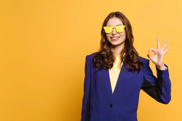 Felice, ragazza elegante in occhiali gialli mostrando gesto ok isolato su giallo — Foto stock