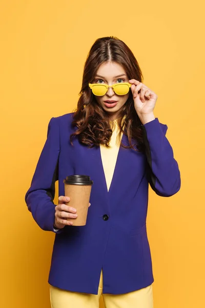 Menina atraente tocando óculos amarelos enquanto olha para a câmera e segurando café para ir isolado no amarelo — Fotografia de Stock