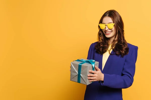 Стильна, усміхнена дівчина в жовтих окулярах тримає подарункову коробку на жовтому тлі — стокове фото