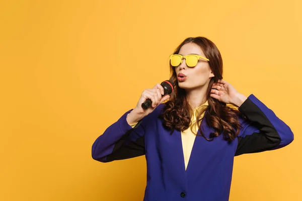 Bela cantora em óculos amarelos cantando com microfone no fundo amarelo — Fotografia de Stock