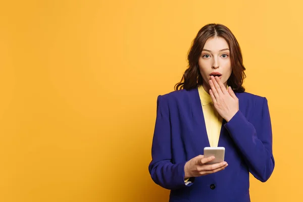 Schockiertes Mädchen bedeckt Mund mit Hand, während es Smartphone auf gelbem Hintergrund hält — Stockfoto