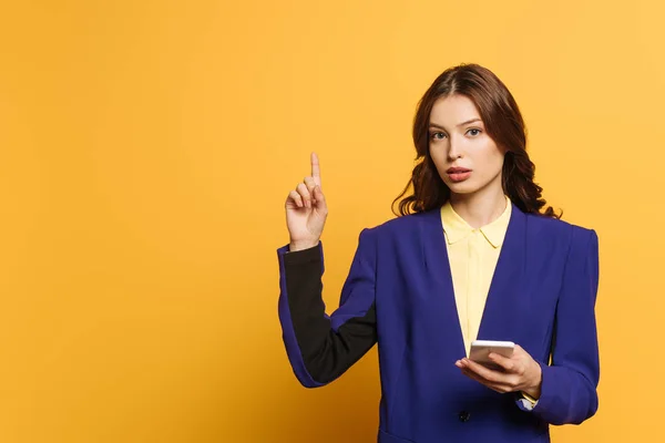 Серьезная девушка держит смартфон и показывает жест идеи на желтом фоне — стоковое фото