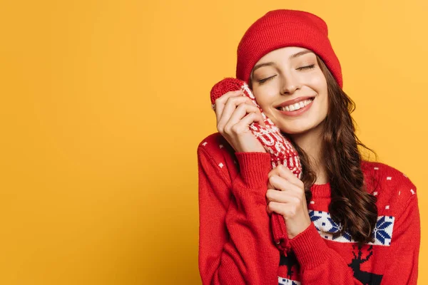 Chica feliz en sombrero y suéter ornamental rojo sosteniendo manoplas cerca de la cara con los ojos cerrados sobre fondo amarillo - foto de stock