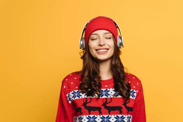 Chica feliz en sombrero y suéter ornamental rojo escuchando música en auriculares inalámbricos con los ojos cerrados aislados en amarillo - foto de stock