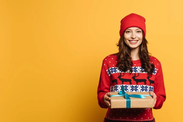 Niña sonriente en sombrero y suéter ornamental rojo sosteniendo caja de regalo sobre fondo amarillo - foto de stock