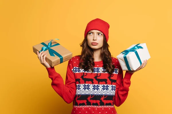 Вдумчивая девушка в шляпе и красный декоративный свитер держа подарочные коробки на желтом фоне — стоковое фото