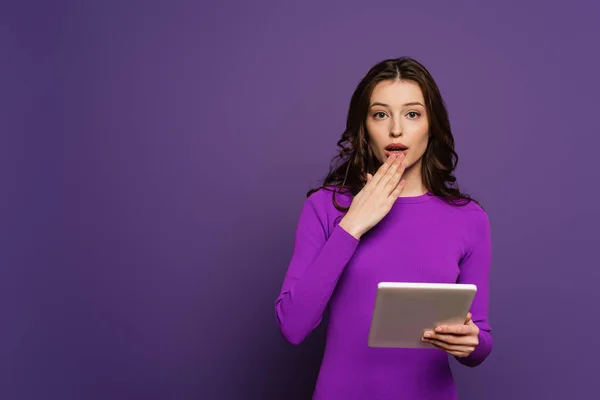 Schockiertes Mädchen, das den offenen Mund mit der Hand bedeckt, während es ein digitales Tablet auf lila Hintergrund hält — Stockfoto