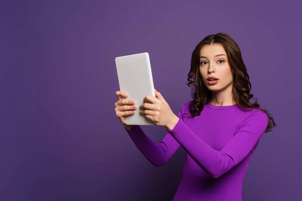 Шокированная девушка смотрит в камеру, держа цифровой планшет на фиолетовом фоне — стоковое фото