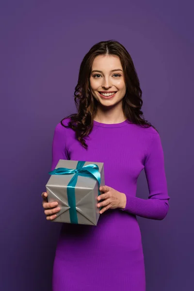 Happy girl holding gift box while smiling at camera on purple background — Stock Photo