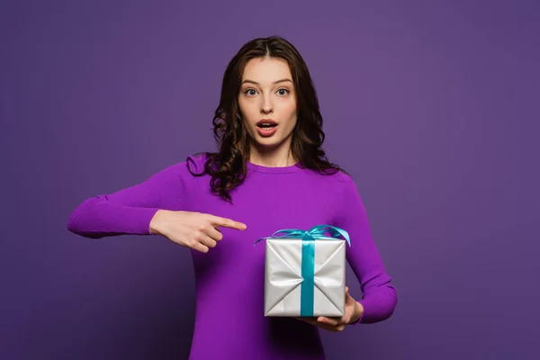 Здивована дівчина вказує пальцем на подарункову коробку на фіолетовому фоні — стокове фото