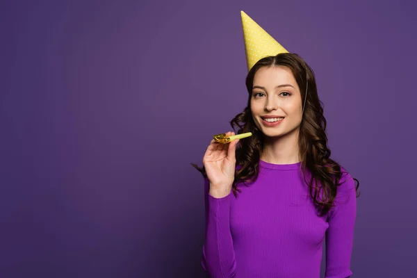 Fille souriante dans le chapeau de fête tenant corne de fête tout en regardant la caméra sur fond violet — Photo de stock