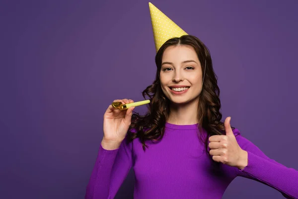 Happy girl in party cap holding party horn and showing thumb up on purple background — Stock Photo