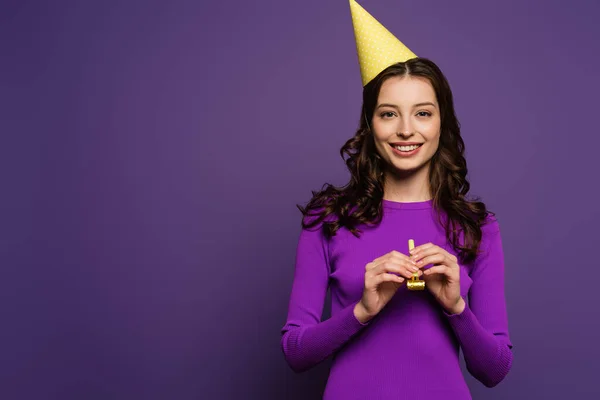 Счастливая девушка в кепке, держащая партийный рог и улыбающаяся камере на фиолетовом фоне — стоковое фото