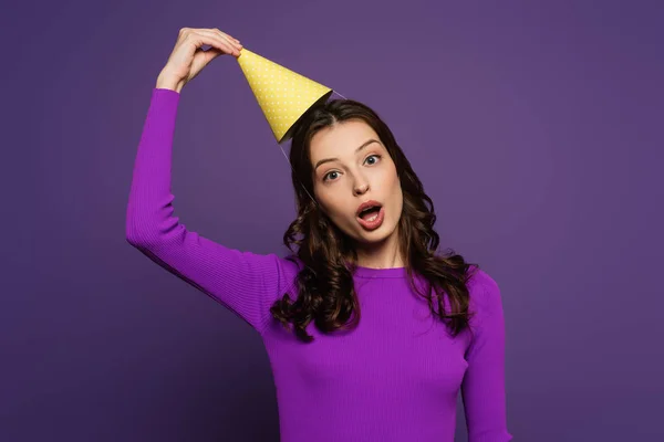 Fille gaie avec bouche ouverte toucher chapeau de fête sur fond violet — Photo de stock