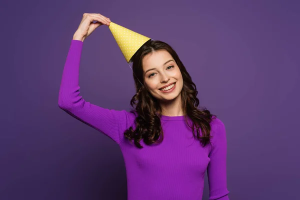 Happy girl touching party cap while smiling at camera on purple background — Stock Photo