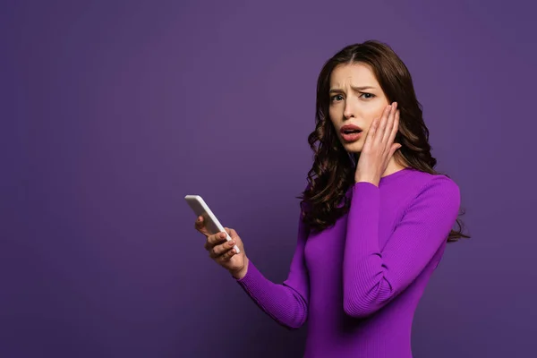 Shocked girl touching face while holding smartphone on purple background — Stock Photo