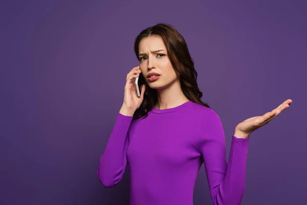 Displeased girl talking on smartphone while standing with open arm on purple background — Stock Photo