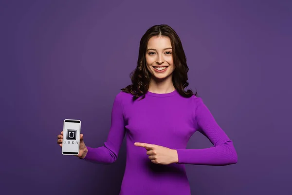 KYIV, UKRAINE - 29 de novembro de 2019: menina sorridente apontando com o dedo para o smartphone com o aplicativo Uber na tela no fundo roxo — Fotografia de Stock