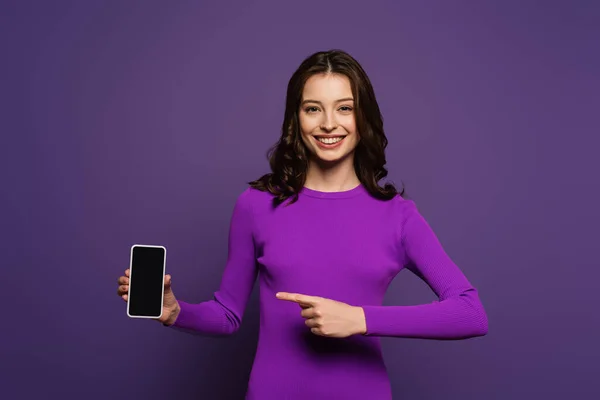 Усмішка дівчинка, що вказує пальцем на смартфон з чистим екраном на пурпуровому фоні — стокове фото