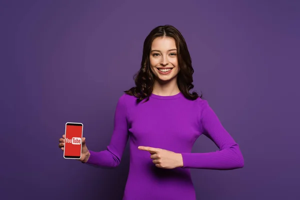 Kiew, Ukraine - 29. November 2019: Lächelndes Mädchen zeigt mit dem Finger auf Smartphone mit Youtube-App auf dem Bildschirm auf lila Hintergrund — Stockfoto