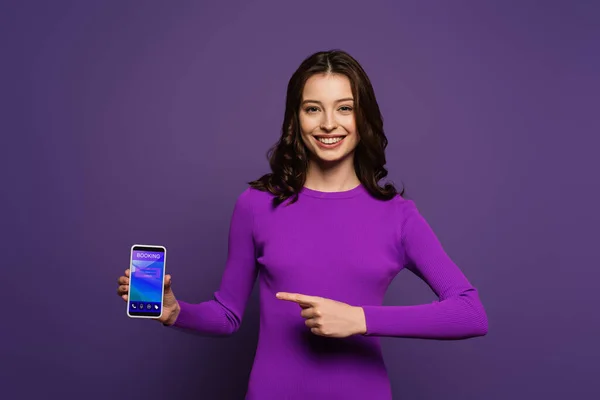 Chica sonriente apuntando con el dedo en el teléfono inteligente con aplicación de reserva en la pantalla sobre fondo púrpura - foto de stock