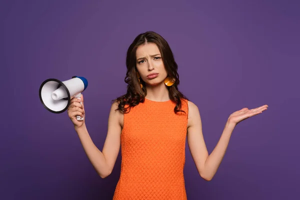 Расстроенная девушка показывает жест пожатия плечами, держа мегафон и глядя на камеру, изолированную на фиолетовый — стоковое фото