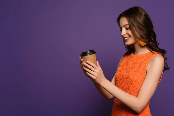 Chica feliz sosteniendo café para ir y sonriendo sobre fondo púrpura — Stock Photo