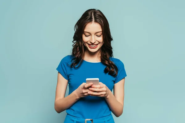 Alegre chica sonriendo mientras chatea en el teléfono inteligente aislado en azul - foto de stock