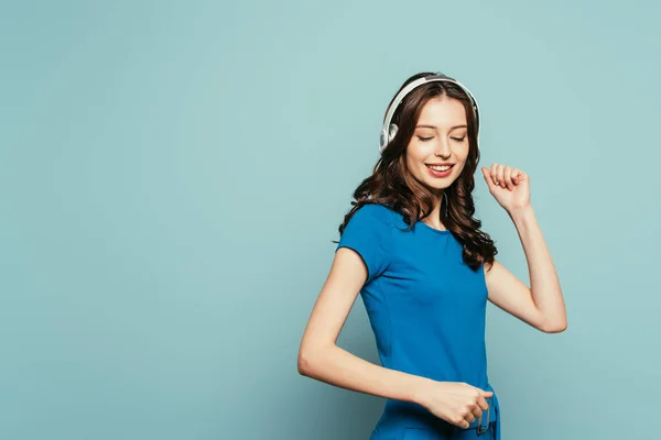 Menina feliz dançando com olhos fechados enquanto ouve música em fones de ouvido sem fio no fundo azul — Fotografia de Stock