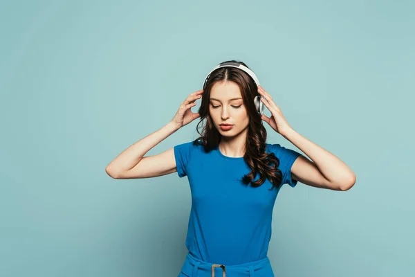 Chica soñadora escuchando música en auriculares inalámbricos con los ojos cerrados sobre fondo azul - foto de stock