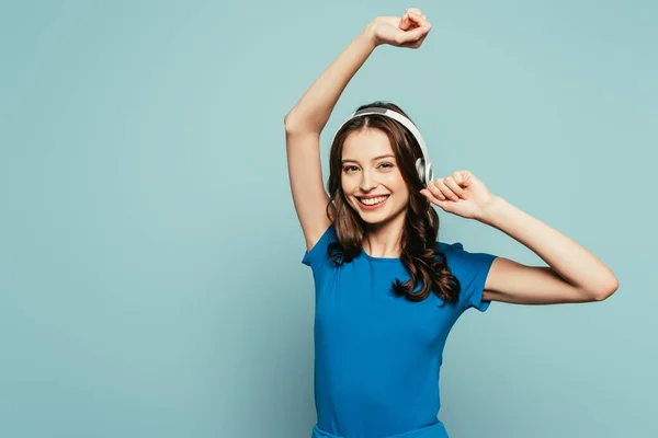 Feliz chica bailando mientras escucha música en auriculares inalámbricos aislados en azul - foto de stock