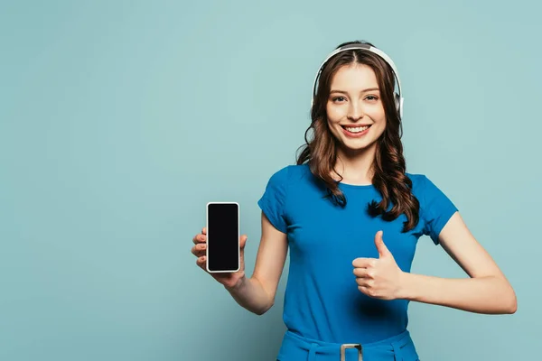 Fille gaie dans les écouteurs sans fil montrant pouce levé tout en tenant smartphone avec écran bancaire sur fond bleu — Photo de stock