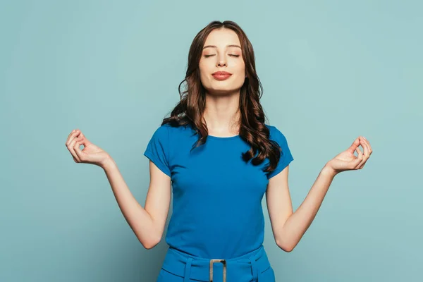 Sonriente chica de pie en la meditación pose con los ojos cerrados sobre fondo azul - foto de stock