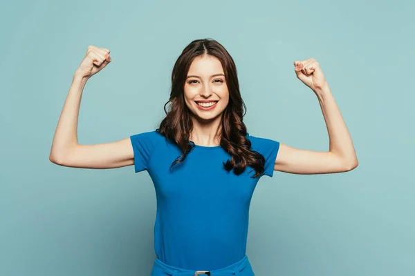 Cheerful girl showing winner gesture while smiling at camera on blue background — Stock Photo