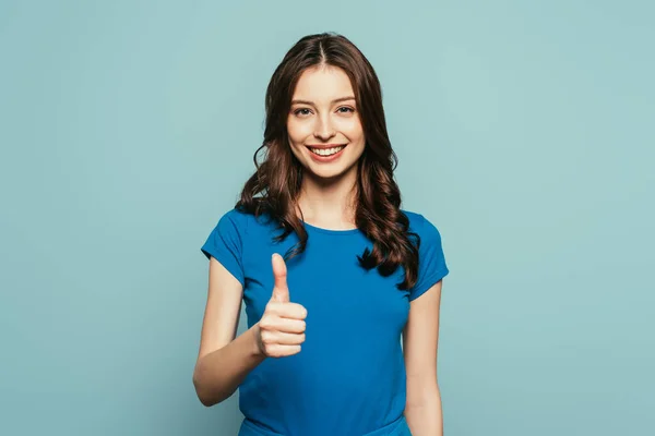 Счастливая девушка, показывающая большой палец вверх, улыбаясь камере на голубом фоне — стоковое фото