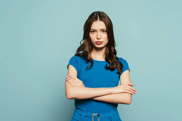 Серьезная, привлекательная девушка смотрит в камеру, стоя со скрещенными руками на синем фоне — стоковое фото