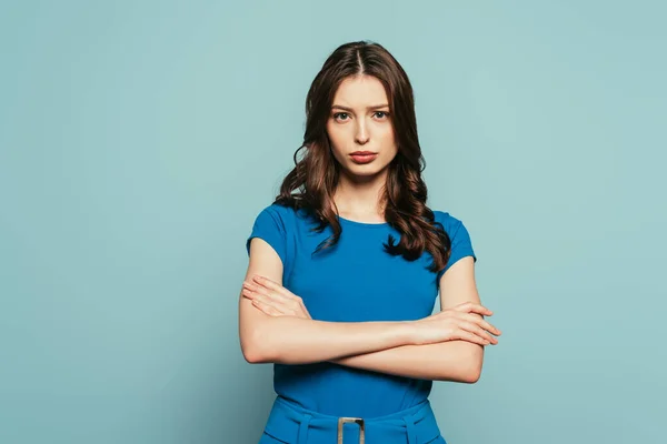 Ernstes, attraktives Mädchen, das mit verschränkten Armen vor blauem Hintergrund in die Kamera blickt — Stockfoto