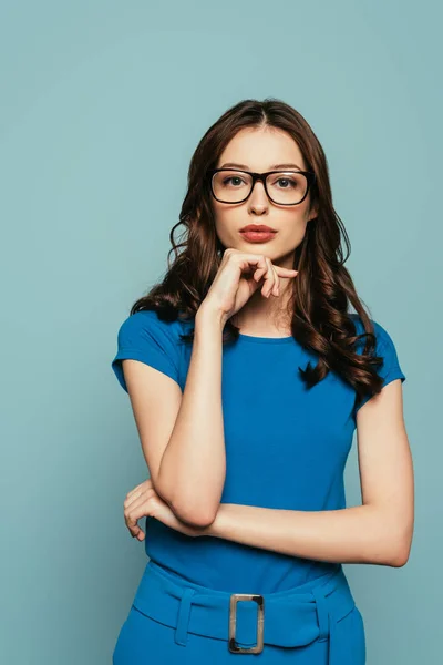 Atractiva, chica confiada mirando a la cámara mientras sostiene la mano cerca de la barbilla aislada en azul - foto de stock