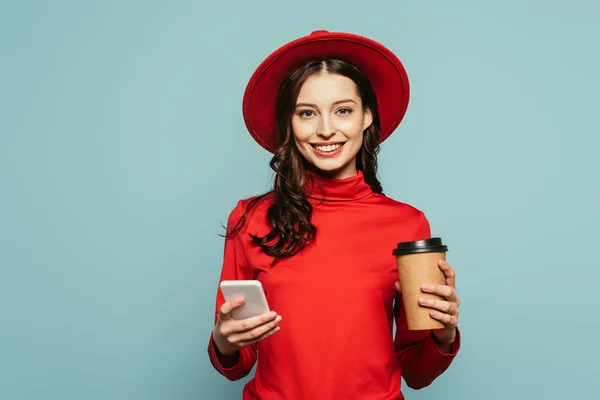 Alegre chica con estilo utilizando el teléfono inteligente mientras sostiene el café para ir aislado en azul - foto de stock