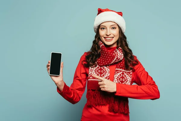Cheerful girl in santa hat and red sweater pointing with finger at smartphone with blank screen on blue background — Stock Photo