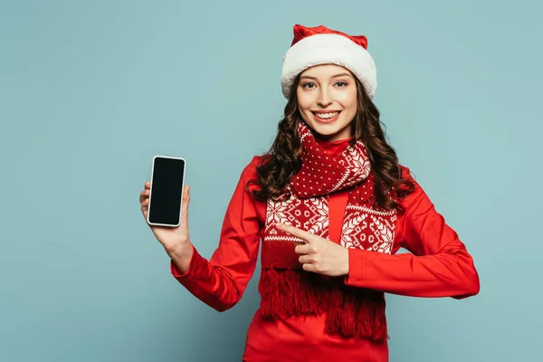 Chica alegre en sombrero de santa y suéter rojo apuntando con el dedo en el teléfono inteligente con pantalla en blanco sobre fondo azul - foto de stock