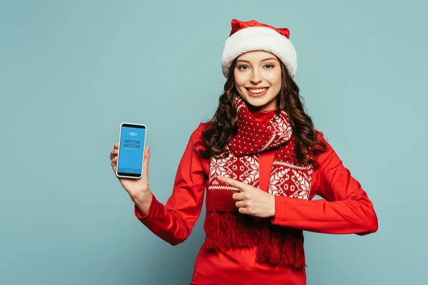 1KYIV, UKRAINE - NOVEMBRO 29, 2019: menina feliz em chapéu de santa e camisola vermelha apontando com o dedo para o smartphone com o aplicativo Skype na tela no fundo azul — Fotografia de Stock