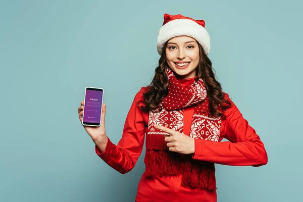 1KYIV, UCRANIA - 29 de noviembre de 2019: chica feliz con sombrero de santa y suéter rojo apuntando con el dedo al teléfono inteligente con la aplicación Instagram en la pantalla sobre fondo azul - foto de stock