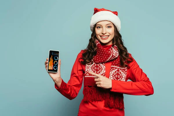 Heureuse fille en santa chapeau et pull rouge pointant du doigt le smartphone avec des graphiques et des graphiques à l'écran sur fond bleu — Photo de stock