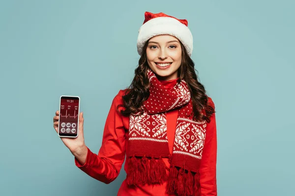 Cheerful girl in santa hat and red sweater showing smartphone with heartbeat rate on screen on blue background — Stock Photo