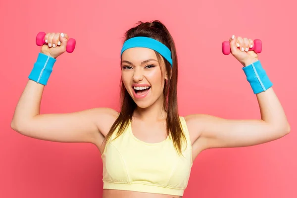 Excited sportswoman training with dumbbells while looking at camera on pink background — Stock Photo